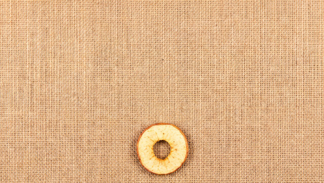 Single dried apple ring placed in the bottom center of a brown woven burlap fabric background