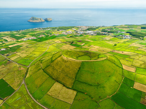 Aerial view of Pico Dona Joana volcanic crater on Terceira Island surrounded by green fields and stone walls with the Atlantic Ocean in the background in Angra do Heroismo, Azores, Portugal.