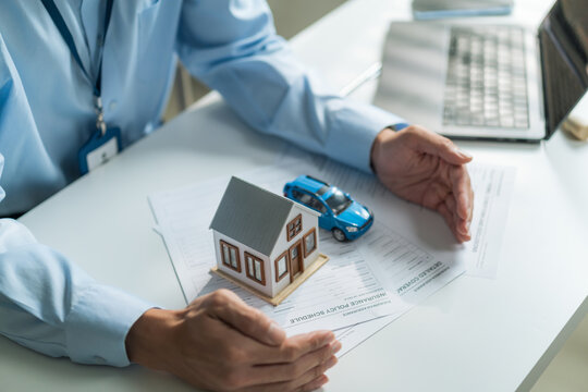 Close-up of hands protecting house model and toy car on insurance policy document for asset protection concept.