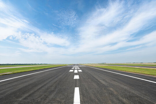 An empty airport runway extends straight into the distance, with clear sky and surrounding grass under sunny weather.