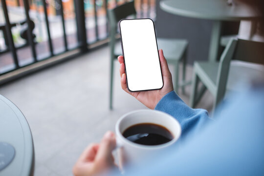 Mockup image of a woman holding and using mobile phone with blank desktop screen while drinking coffee in cafe