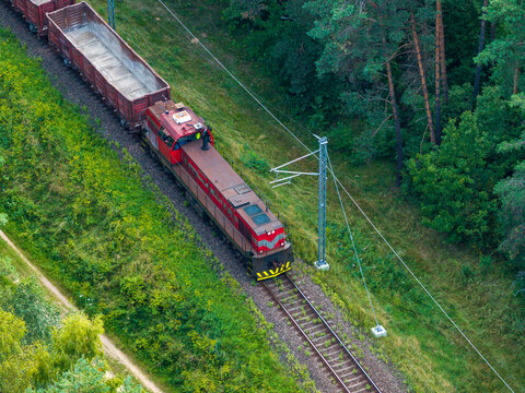 Aerial view of a red freight train locomotive pulling cargo wagons along tracks through a lush green forest in Vilnius, Vilnius County, Lithuania.