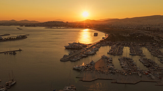 Aerial view of a marina filled with yachts and boats at sunset with the golden sun reflecting on the calm water and the city skyline in Athens, Greece.