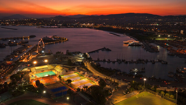 Aerial view of a marina with illuminated sports courts, including a swimming pool and tennis courts, situated along the coastline of Athens, Greece.