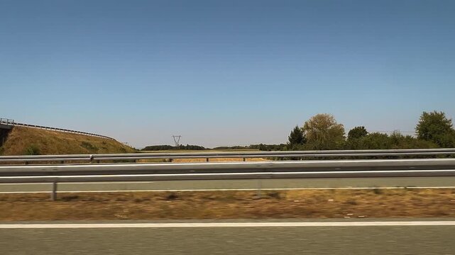 Wide shot taken from a moving vehicle on a highway looking up at a concrete overpass bridge supported by heavy pillars, with metal guardrails running alongside the road, 4K.