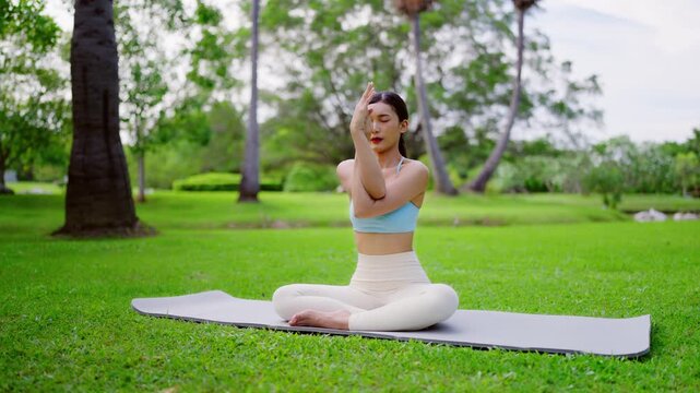 Beautiful young Asian woman practicing yoga outdoors on sunny day at public park.Serene female peaceful and mindfulness sit on exercise mat make lotus pose
