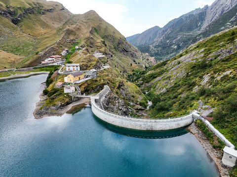 Aerial view of the Diga e Lago di Malciaussia with its curved dam wall, surrounding green mountain slopes, and small buildings nestled on the hillside in Usseglio, Piedmont, Italy.