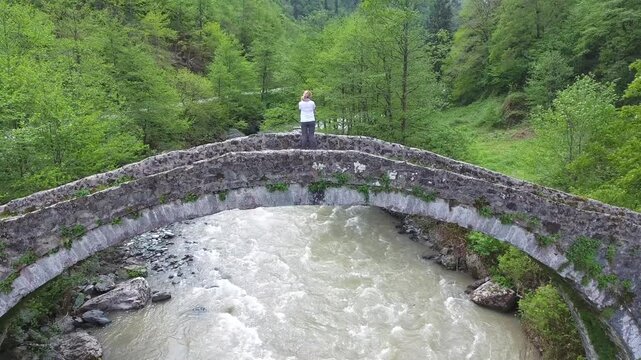 Blonde woman crosses historic stone arch bridge above forest river in Rize Turkey.