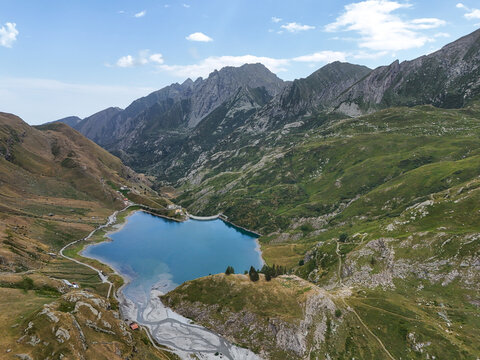 Aerial view of Diga e Lago di Malciaussia surrounded by rugged alpine mountains, green slopes, and rocky terrain under a bright blue sky in Usseglio, Piedmont, Italy.