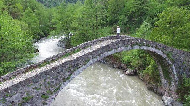 Blonde woman crosses historic stone arch bridge above forest river in Rize Turkey.