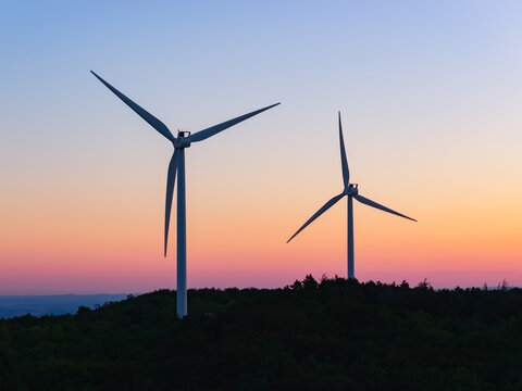 Aerial view of two wind turbines rising above a forested landscape against a colorful sunset sky, highlighting renewable energy in Marsanne, Auvergne-Rhone-Alpes, France.
