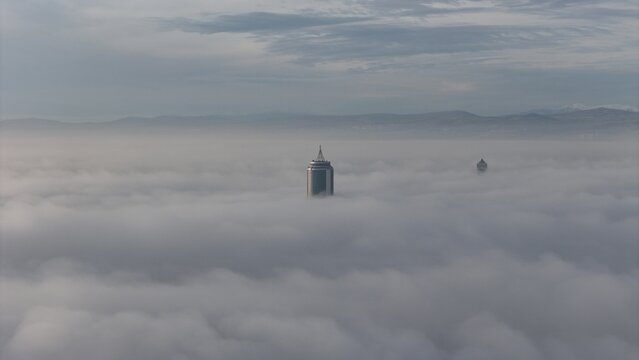 Aerial view of Selcuk Tower rising above a thick layer of morning fog with distant mountains visible under a cloudy sky in Konya, Konya, Turkiye.