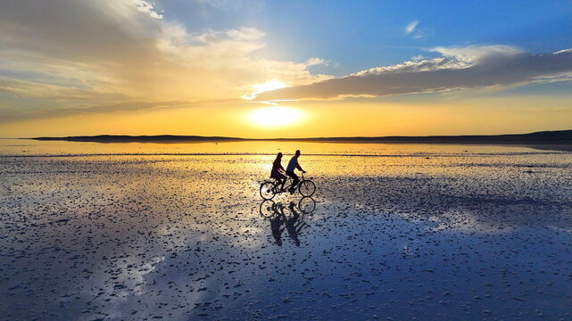 Aerial view of a couple riding a tandem bicycle across the reflective salt flats of Tuz Golu at sunset in Sereflikochisar, Ankara, Turkiye.