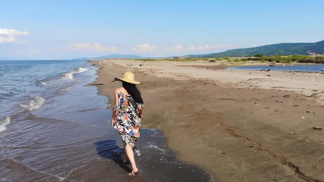 Barefoot woman walks along sandy beach with colorful skirt, sea waves calm summer atmosphere.