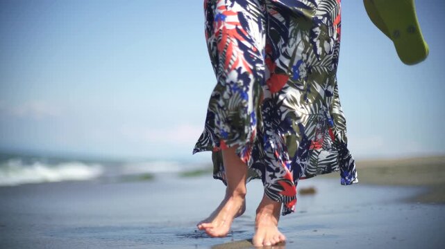 Close view of woman feet walking on beach with sea waves, wet sand flowing skirt.