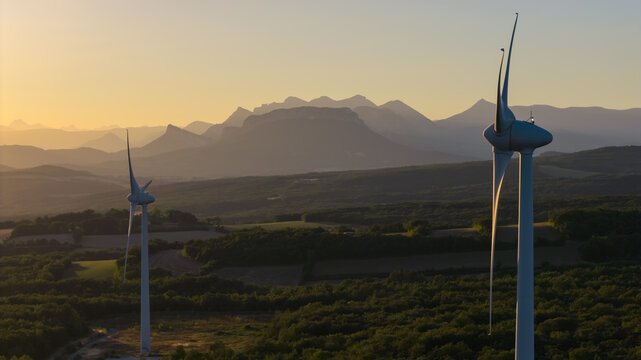 Aerial view of wind turbines rising above a rural landscape with dramatic mountain ranges in the background during golden morning light in Marsanne, Auvergne-Rhone-Alpes, France.