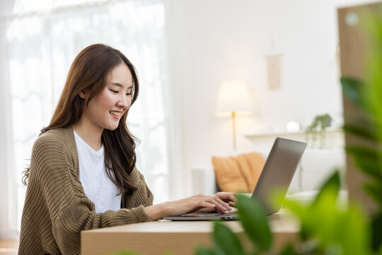 Woman laptop home office typing smiling, young Asian freelancer working at wooden desk in bright living room with plant, calm remote job lifestyle