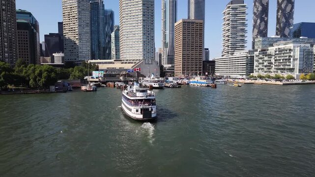 High-angle stationary shot looking down at a double-decker passenger ferry boat departing the Jack Layton Ferry Terminal, leaving a white water wake with the Toronto skyline in the background