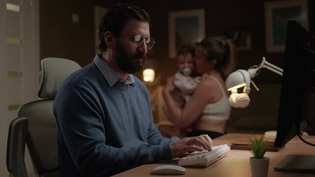 Man with glasses working at computer in home office at night. Wife and child on background. Online work concept