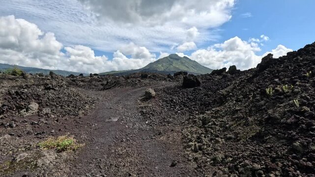 Wide shot of a black solidified lava valley with a dirt path and a large active volcano on the horizon under blue sky and white clouds on Bali island during daytime.