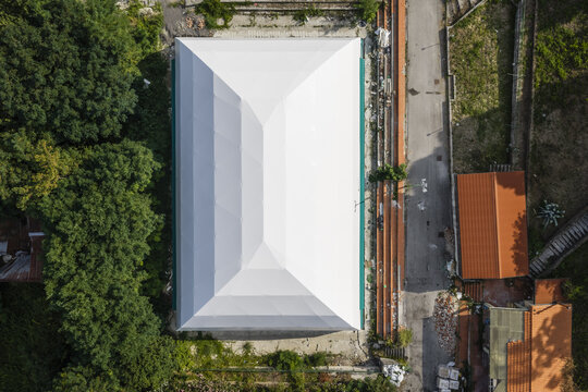 Aerial view of a rectangular building with a white roof, surrounded by green trees, a road, and structures with terracotta roofs in Summonte, Campania, Italy.