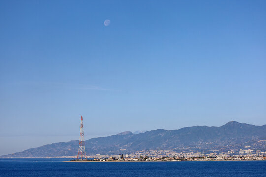 The historic red and white steel electricity Pylon of Messina at Torre Faro under a daytime moon, Sicily.