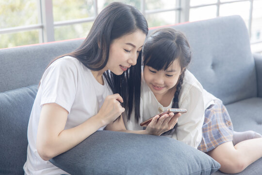 Happy mother and daughter using smartphone together on sofa at home with natural light