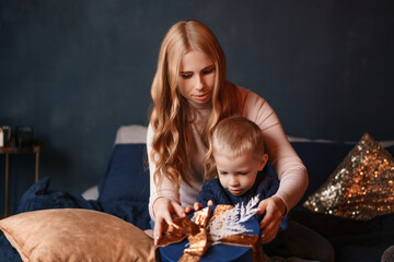 Naklejka premium Hands of a young caucasian mother and her little son unwrapping a blue present with a golden bow at home, festive surprise image for christmas gift campaigns.