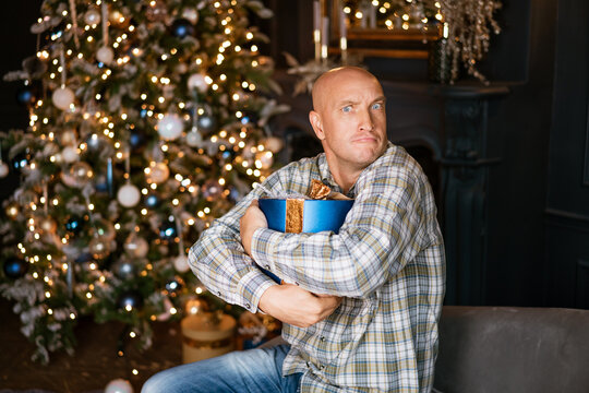 Mature Caucasian man in a plaid shirt clutches a blue gift box with a comic reluctant grimace by the Christmas tree - festive humor content for seasonal campaigns and cards.