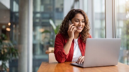 Obraz premium Young woman with curly brown hair sitting at a desk talking on a smartphone with a laptop open in front