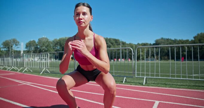 Young athletic girl in sportswear preparing for training at the stadium, warming up and jumping on the running tracks on a sunny summer day