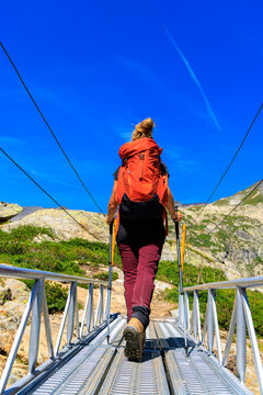 Rear view of female hiker with red backpack crossing metal suspension bridge in alpine mountains