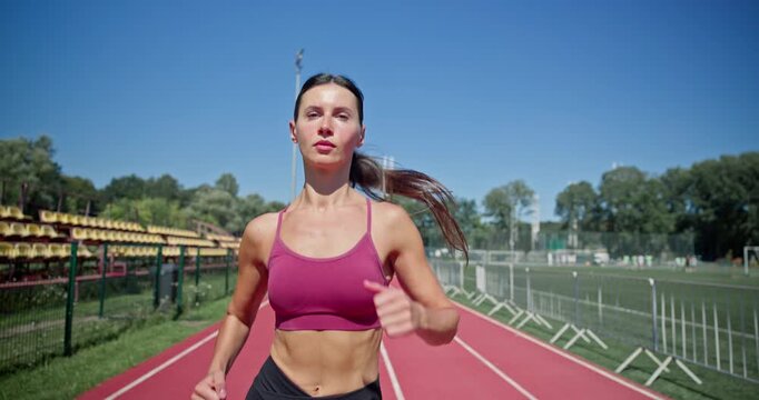 Young athletic girl in sportswear preparing for training at the stadium, warming up and running on the running tracks on a sunny summer day