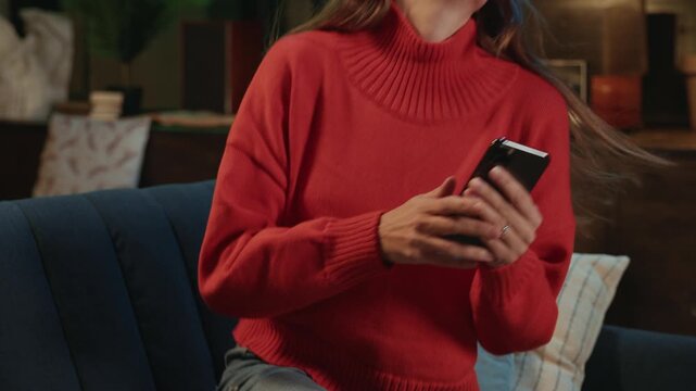 Back shot of slim young woman in blue jeans and coral sweater taking smartphone and sitting on couch. Cute smiling Caucasian girl resting on sofa looking on screen of gadget
