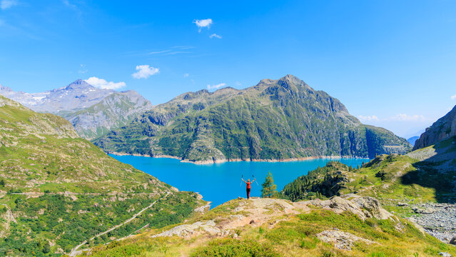 Happy female hiker standing with arms raised overlooking turquoise alpine lake and mountain range. Emosson Lake in Switzerland