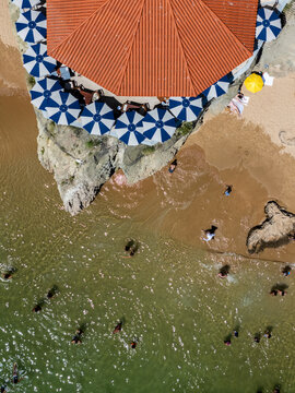 Aerial view of a restaurant with blue and white umbrellas on a rocky outcrop overlooking a sandy beach with people swimming in the clear water in Cascais, Lisbon, Portugal.