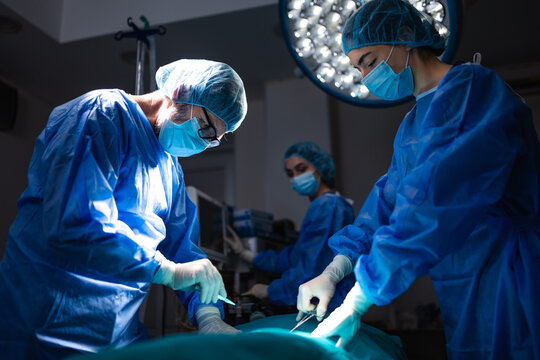 Surgeon focused on operation while female doctors assist in hospital operating room. Medical staff wearing surgical scrubs and masks during medical procedure under bright surgical lights overhead.