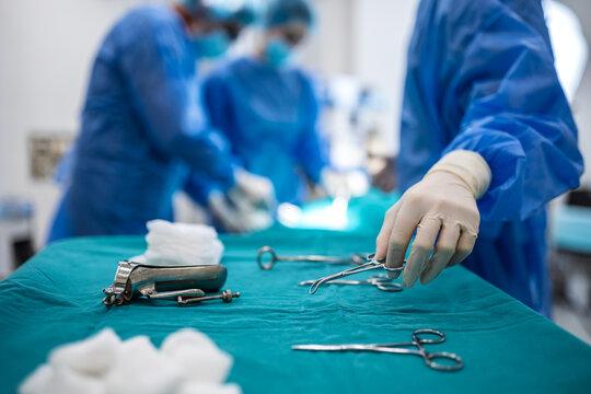 Gloved hand picking up surgical instrument from tray in hospital operating room. Medical equipment and tools prepared for surgery procedure with surgical team working in background.