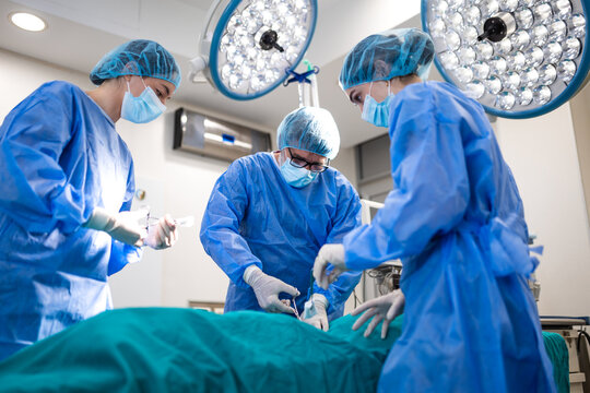 Surgeon and female doctors performing surgery in hospital operating room. Medical team wearing surgical scrubs and masks during successful operation under bright surgical lights.