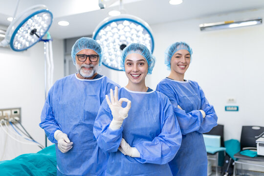 Surgeon and female doctors smiling in hospital operating room. Medical team wearing surgical scrubs and masks posing together after successful operation under bright surgical lights.
