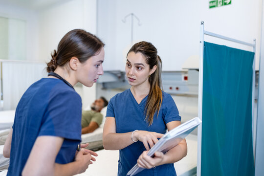 Two female doctors discussing medical chart in hospital ward. Healthcare professionals in blue scrubs reviewing patient records with elderly man resting in hospital bed in background.