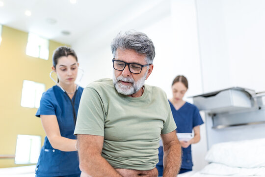 Senior man sitting on hospital bed holding back in pain with female doctor examining him. Elderly patient receiving medical care and physical examination from healthcare professionals.
