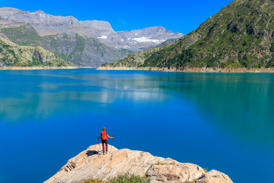 Female hiker with red backpack standing on rock edge overlooking vivid blue alpine lake and mountains- Freedom, adventure, wanderlust concept