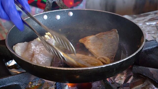 A chef in a restaurant kitchen is frying fish in a hot pan. The fish sizzles as it cooks on the heated surface.