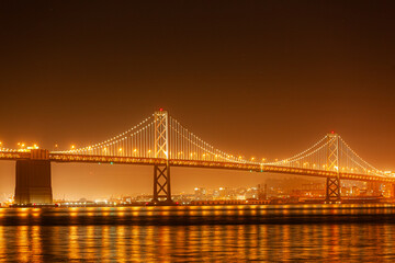 Fototapeta premium The illuminated San Francisco-Oakland Bay Bridge glows at night over the water in San Francisco, California, USA, reflecting the city lights.
