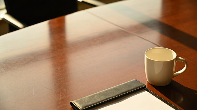 Glass cup of hot tea with infuser and yellow notepad on wooden conference table in sunny office