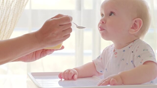 Slow motion: feeding a baby from a spoon. Mom feeds a 9 month baby with fruit puree from a spoon, close-up, high key. Meal time. Little boy eats sitting on baby's chair. 