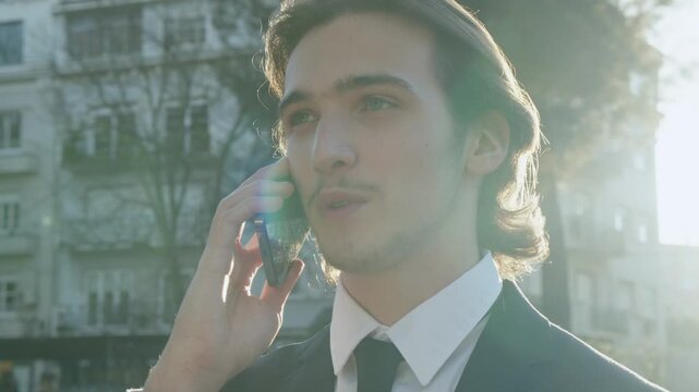 Smiling Man in a business suit stands outdoors and talking on the phone. A young businessman in a suit talking on a smartphone. A smiling businessman in the city talking on a cell phone. 