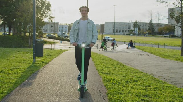 Young smiling man rides an electric scooter in the city on a sunny day. Happy student riding on a rented green electric scooter through the streets of the city. Ecological transport. Slow motion