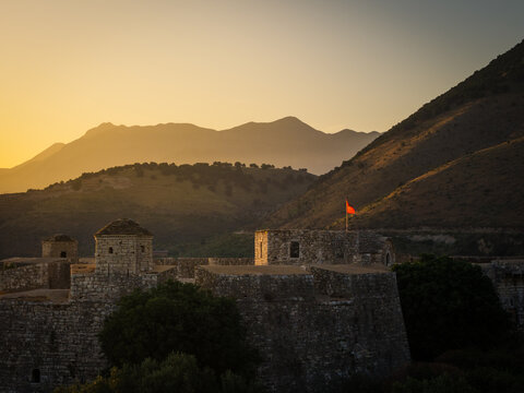 Aerial view of the historic Porto Palermo Castle with stone walls and towers glowing in the golden hour light, set against rolling mountains in Porto Palermo, Qarku i Vlores, Albania.